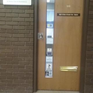 A wooden office door with a vertical window and signs, set in a brick wall.