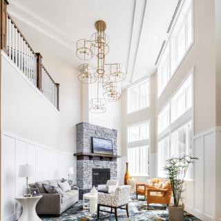 A spacious living room with a high ceiling, large windows, and a modern design featuring a stone fireplace and elegant chandeliers.