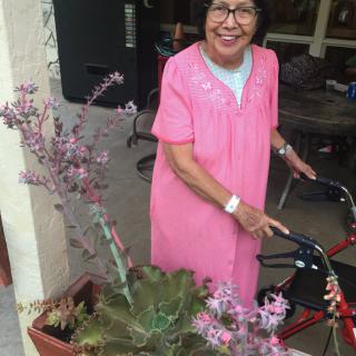 An elderly woman in a pink dress smiles while using a walker outside, surrounded by potted plants.