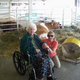 An elderly person in a wheelchair and another person posing by cows inside a farm setting.