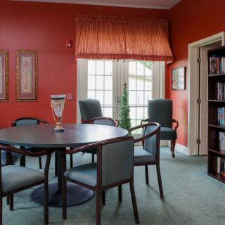 A dining room with red walls, featuring a round table surrounded by chairs, a bookshelf, and large windows with curtains.