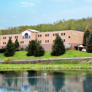 A serene view of a multi-story building alongside a calm body of water, surrounded by lush greenery.