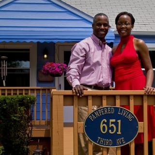 A couple standing on a porch in front of their home, smiling at the camera.