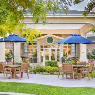 A charming outdoor seating area with tables, chairs, and blue umbrellas set in front of a building surrounded by lush greenery.