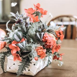 A vibrant floral arrangement with orange flowers in a woven basket sits on a wooden table.
