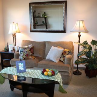 A cozy living room with a sofa, decorative pillows, and a coffee table adorned with fruit and books. Two lamps flank the sofa, adding warmth to the space, while a mirror above reflects the decor.