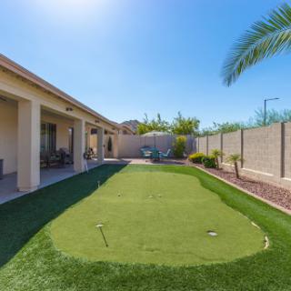 A beautifully designed backyard featuring a golf putting green, surrounded by lush artificial grass and palm trees under a clear blue sky.