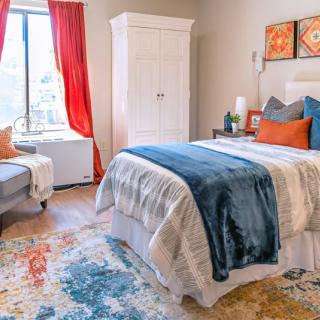 A cozy bedroom featuring a neatly made bed with colorful pillows, an armchair by the window with red curtains, a white wardrobe, and a vibrant rug on the floor.