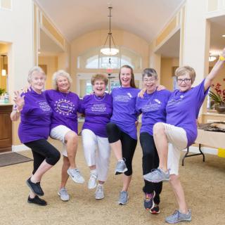 A group of senior women having fun and exercising together indoors, all wearing matching purple shirts.
