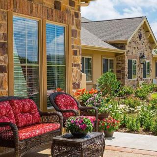 Cozy patio area with wicker furniture and colorful cushions in front of a stone house, surrounded by a lush garden.