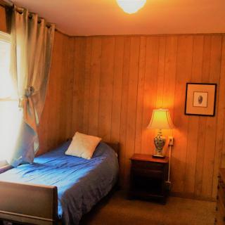 A cozy bedroom with wood-paneled walls, featuring a single bed, nightstand, and a lamp near the window.