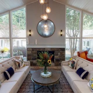 A modern living room with two sofas facing a central coffee table, decorated with floral arrangements. Large windows and a fireplace enhance the bright and cozy atmosphere.