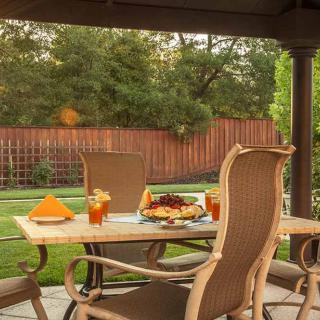 A cozy outdoor patio setup with a table and four chairs, featuring food and drinks, in a lush backyard garden.