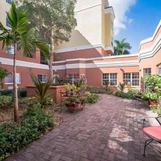 A peaceful courtyard with a brick pathway, surrounded by lush plants and a seating area, adjacent to a residential building on a sunny day.