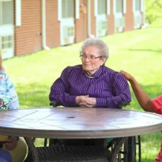 An elderly woman in a wheelchair sits at an outdoor table with companions.
