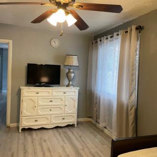 A cozy bedroom features a ceiling fan, dresser with a TV and lamp, and curtains covering a window.
