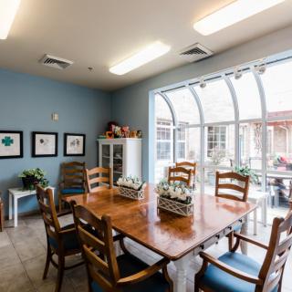 A well-lit dining area with a wooden table and chairs, featuring large windows allowing natural light and decorated with plants.