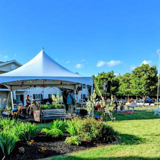 A vibrant outdoor event setup under a large tent, surrounded by lush greenery and clear blue skies.