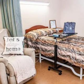A cozy room with a hospital bed and an overbed table, featuring an armchair with a decorative pillow.