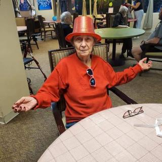 An elderly person wearing a festive red party hat and sweater, sitting at a table indoors, with glasses resting on the table.