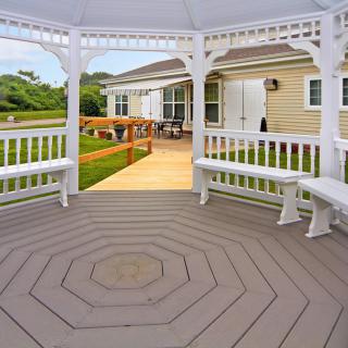 A view from inside a white gazebo with benches, overlooking a wooden deck leading to a house and garden area.