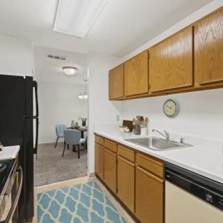 A modern kitchen with wooden cabinets, white countertops, and black appliances, leading into a cozy dining area.