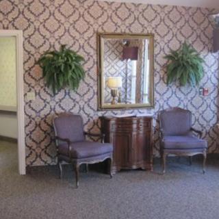 A cozy waiting area with patterned wallpaper, two chairs, a mirror, and decorative plants.