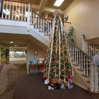A large Christmas tree decorated with lights and ornaments stands by a staircase in a warmly lit interior.