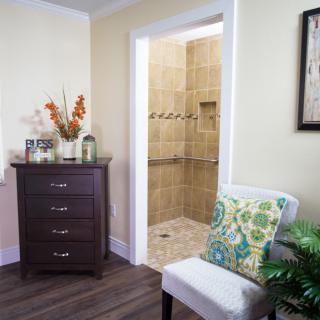 A cozy living space featuring a wooden drawer and comfortable chair with a floral pillow, leading into a tiled bathroom.