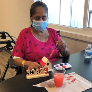 A person wearing a mask is painting a birdhouse at a table.