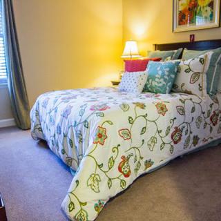 A cozy bedroom with a floral-patterned comforter and decorative pillows, softly lit by a bedside lamp.
