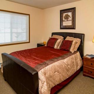 A cozy bedroom featuring a neatly made bed with red and beige bedding, a window, and a nightstand with a lamp.