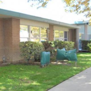 A small, single-story school building with bushes in front and a sidewalk leading to the entrance.
