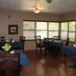 A cozy dining room with tables covered in blue tablecloths, surrounded by wooden chairs and illuminated by natural light from large windows.