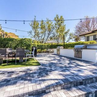 A spacious backyard patio with a dining area and grill, adorned with string lights on a sunny day.