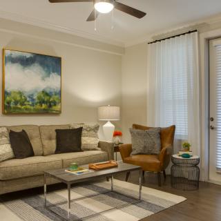 A cozy living room with a sofa, armchair, and coffee table beneath a ceiling fan, decorated with a colorful painting.