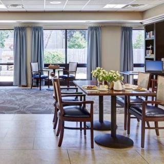 A cozy dining area with tables and chairs, featuring large windows and decorative plants.