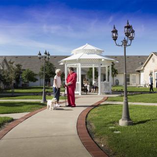 Two people walking with a dog near a gazebo on a sunny day in a nursing home garden.