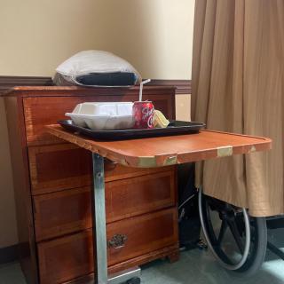 A hospital room scene with a meal tray on an overbed table next to a wheelchair.