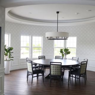 A modern dining room with a large round table and chandelier, featuring wooden flooring and natural light from multiple windows.