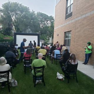 An outdoor event with an audience seated in front of a screen near a brick building.