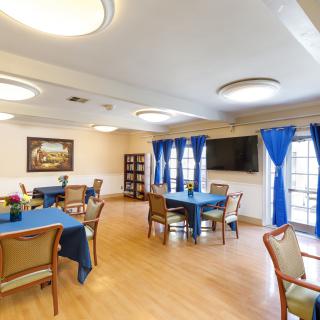 A well-lit dining area with tables and chairs, featuring blue curtains and a television on the wall.