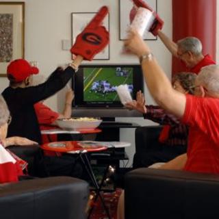 A group of sports fans excitedly watch a football game on TV, wearing red and cheering with foam fingers.