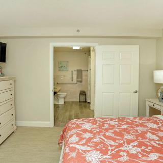 A cozy bedroom with a coral patterned bedspread, featuring a dresser and TV. The room opens to an adjoining bathroom.