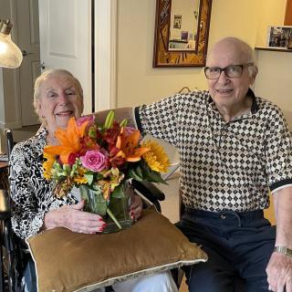 An elderly couple smiling together indoors, holding a colorful bouquet of flowers.