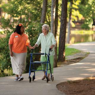 An elderly person using a walker is assisted by a caregiver on a scenic outdoor path.