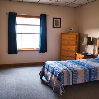 A cozy bedroom featuring a neatly made bed with a plaid blanket, a wooden dresser, and bright blue curtains framing a window.