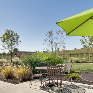 A sunny patio with a table, chairs, and a green umbrella overlooking a landscaped yard.