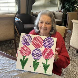 An elderly woman proudly displays her colorful painting of flowers.
