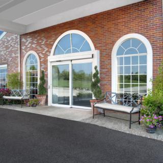 Entrance to a brick building with arched windows and glass doors, featuring benches and potted plants.
